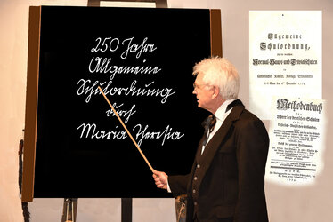 Ein mit historischem Küstüm als Lehrer verkleideter Mann steht mit erhobenem Zeigestab vor einer Schultafel mit der Aufschrift in Kurrent 250 Jahre Allgemeine Schulordnung durch Maria Theresia | © Wolfgang Hochreiter
